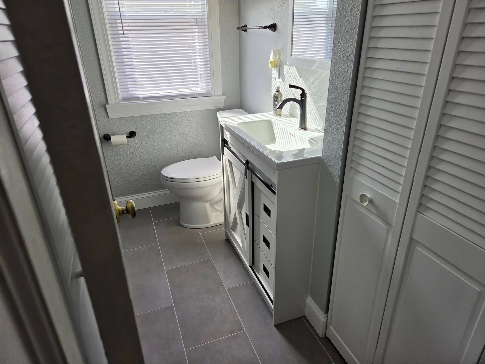 Renovated bathroom with gray tile floor, white vanity, black faucet, and louvered doors