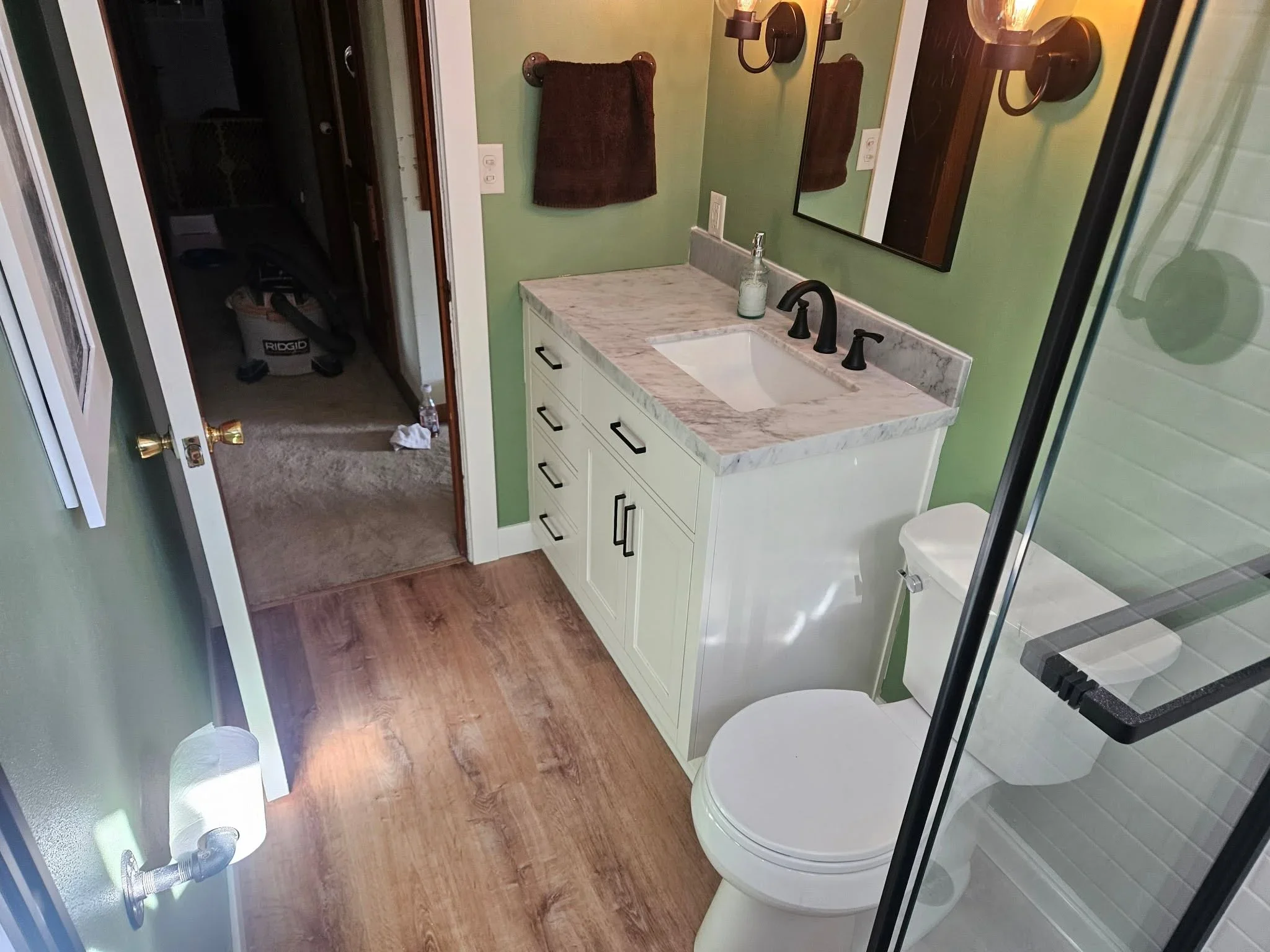White vanity with marble countertop, bronze sconces, sage green walls, wood flooring, and tiled shower
