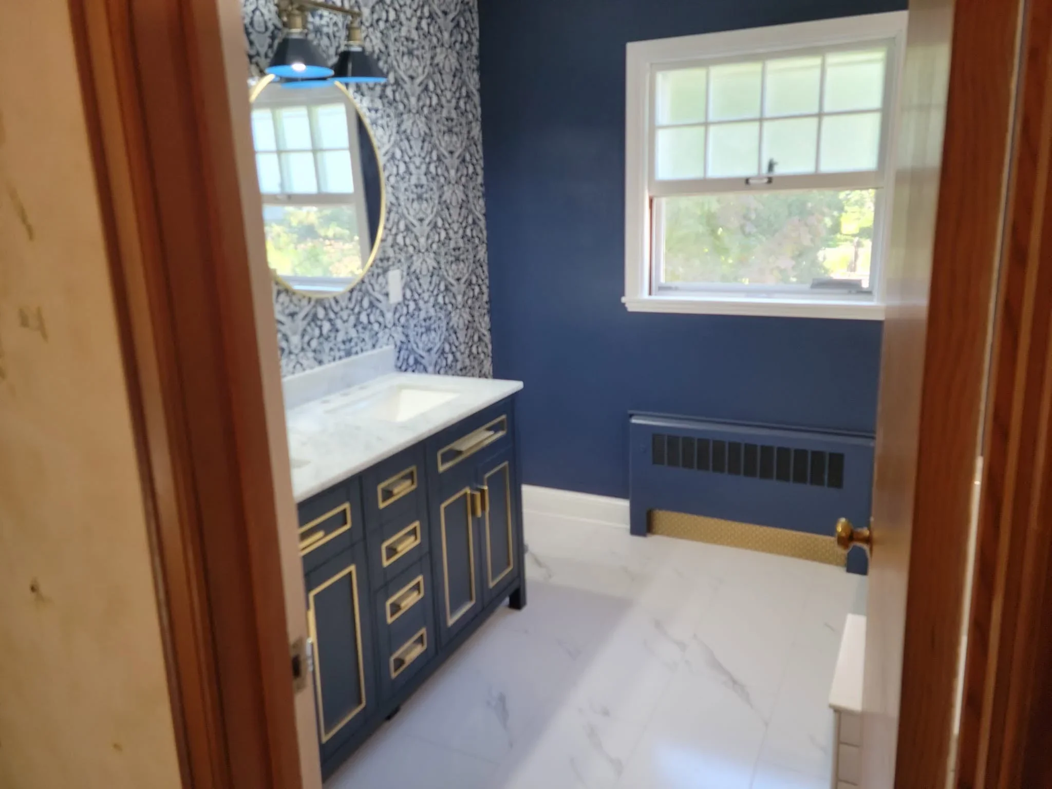 Navy vanity with brass hardware, marble countertop, textured wallpaper, and white tile flooring in renovated bathroom