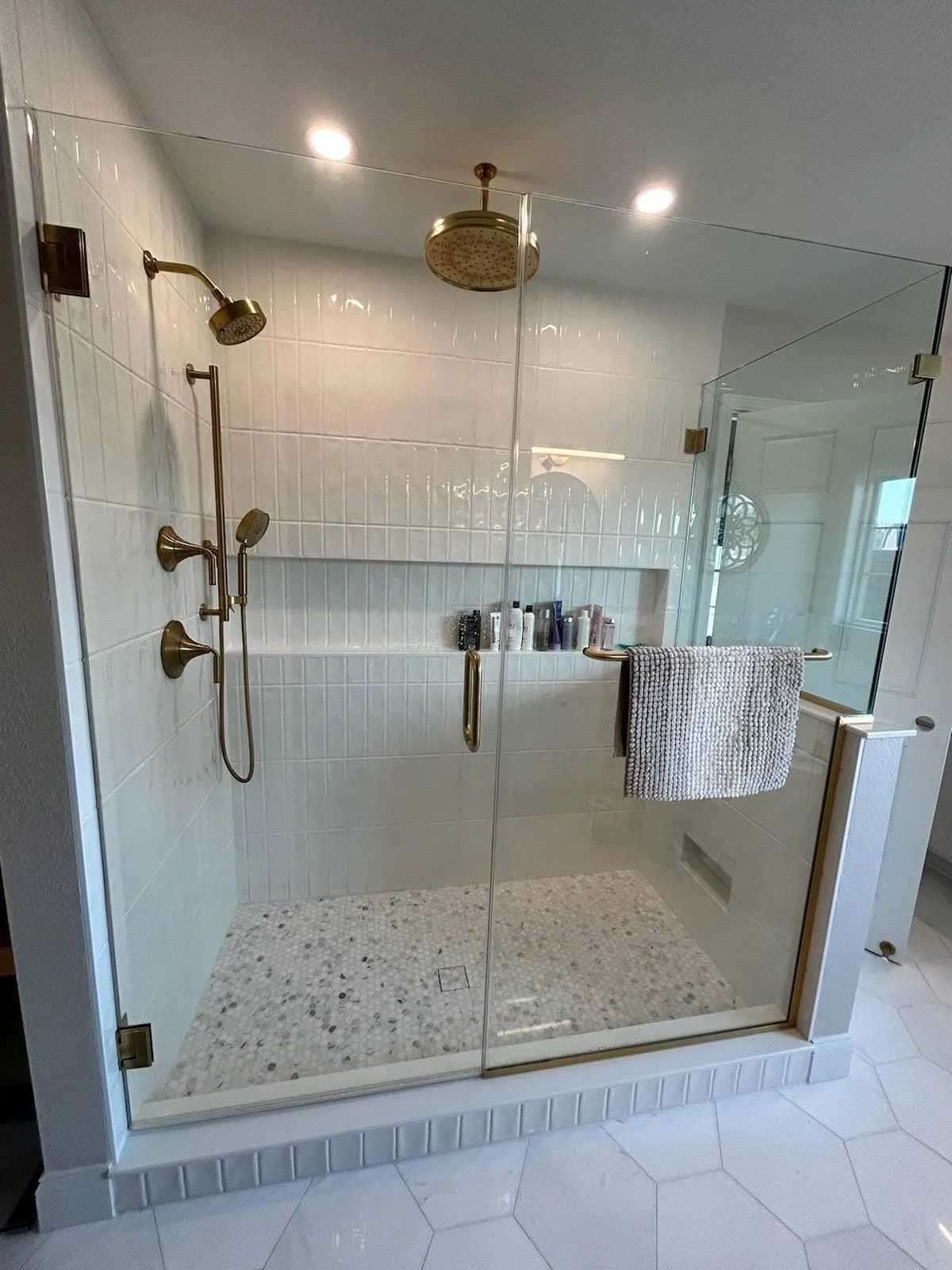 Frameless glass shower with white subway tile, terrazzo floor, dual brass showerheads, and built-in shelving