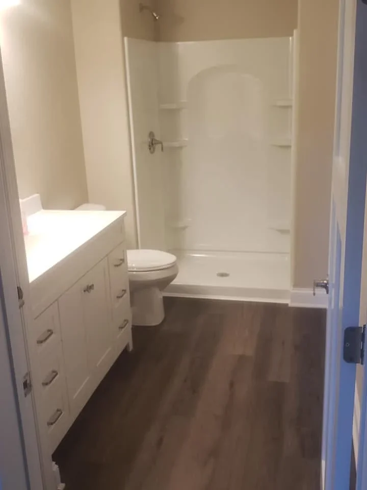 White vanity, toilet, and fiberglass shower surround with dark wood flooring in renovated bathroom