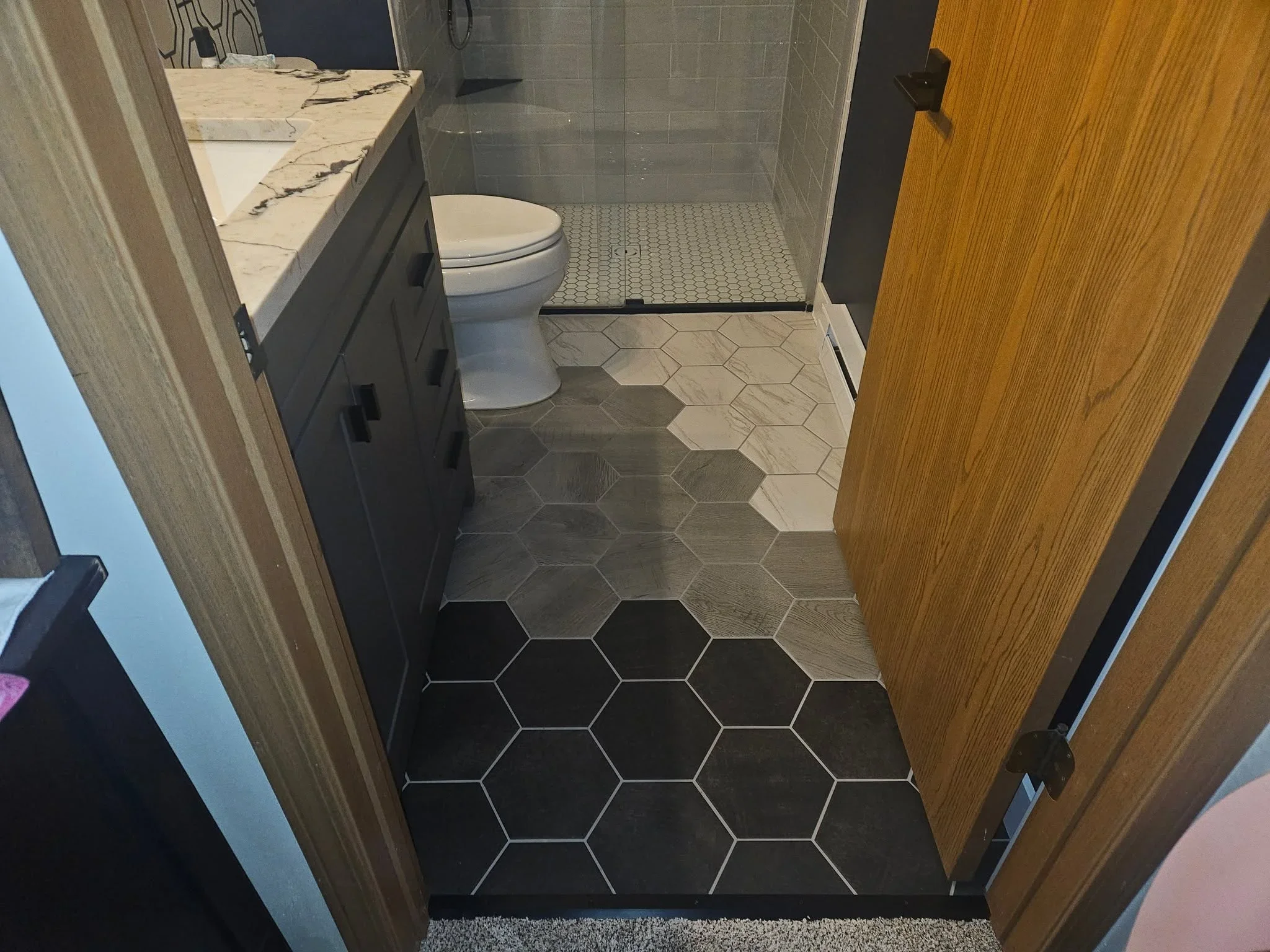 Black hexagonal floor tile with white grout transitioning to white penny tile in shower area, marble vanity countertop, and gray subway tile walls