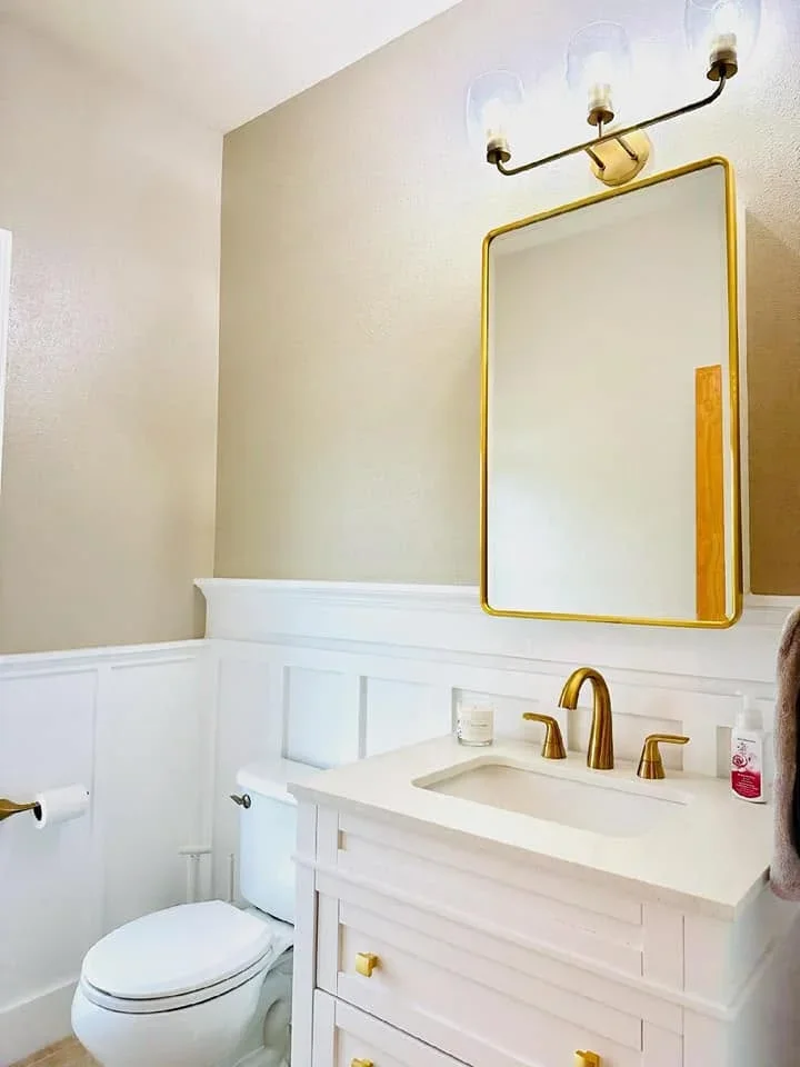 White bathroom vanity with brass faucet, brass-framed mirror, and three-light vanity sconce