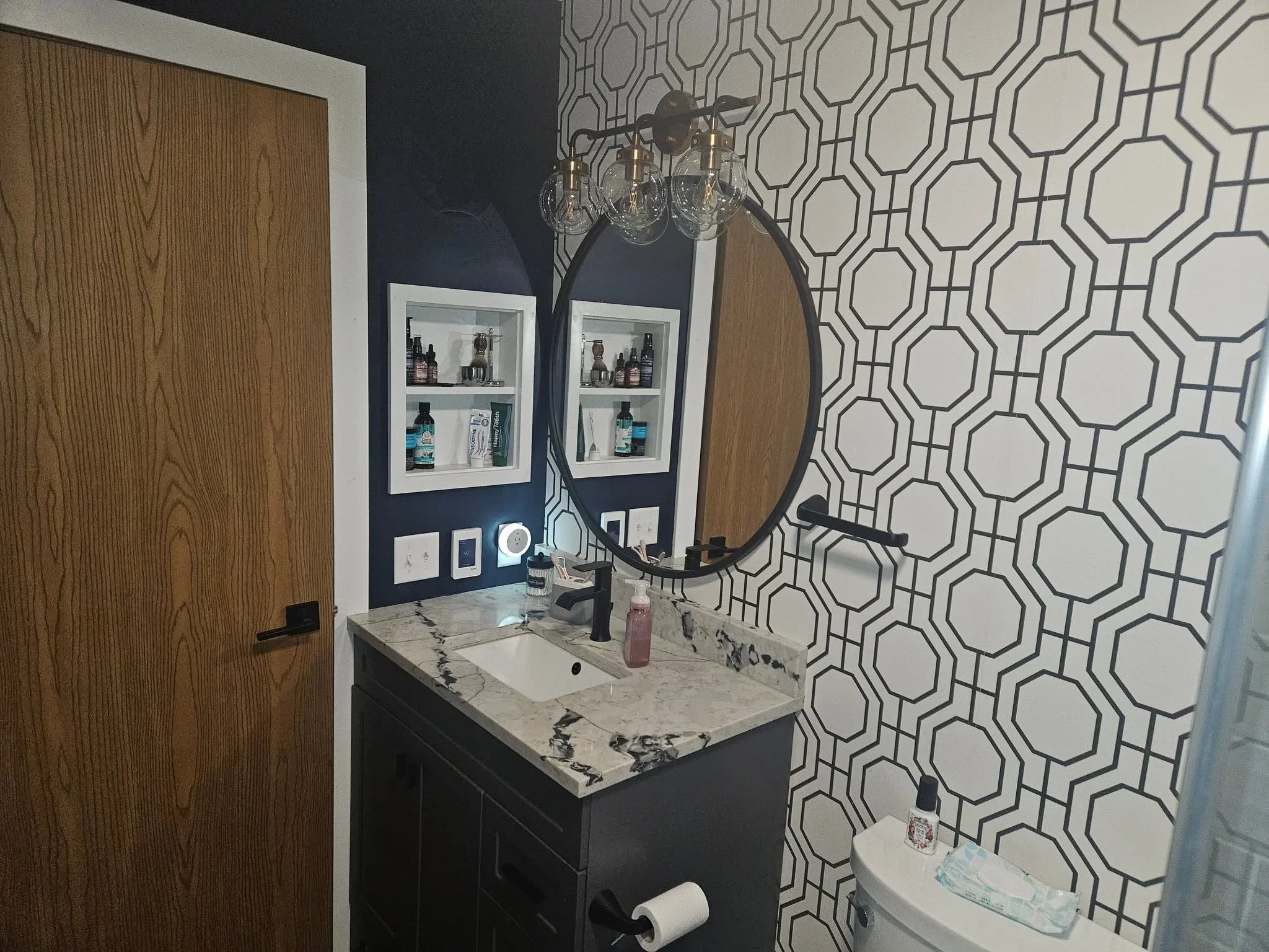 Dark vanity with marble countertop, geometric hexagon tile wall, round mirror, and brass lighting fixture