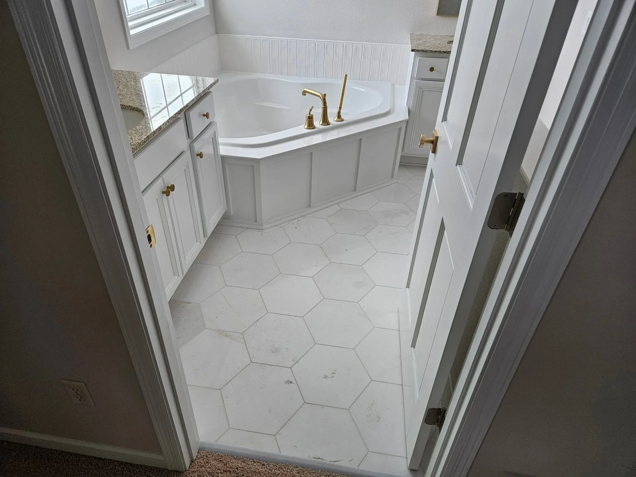 White bathroom with soaking tub, shaker cabinets, hexagonal marble floor tile, and brass fixtures